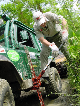 A man with gloves pushes down on the lever of his red Hi-Lift jack to lift his overland vehicle from a muddy puddle.