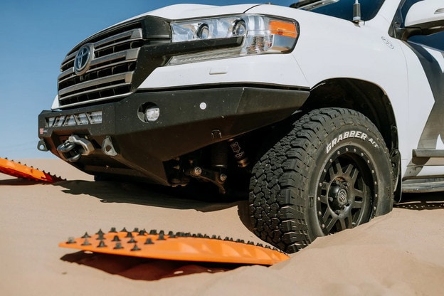 Neon orange recovery tracks with black spikes lie under an overlanding 4x4's wheels that are embedded in the sandy desert with a clear blue sky above. 