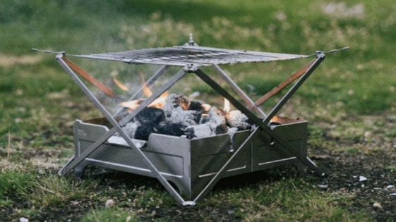 Small portable silver fire pit on the grass with open flames.