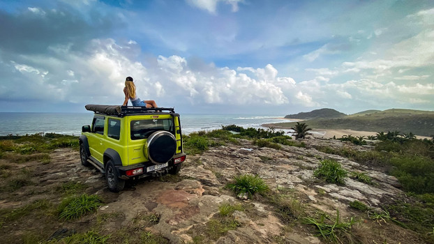 Woman sat on top of a bright green jeep that's parked on top of a cliff, looking over the views of the sea.