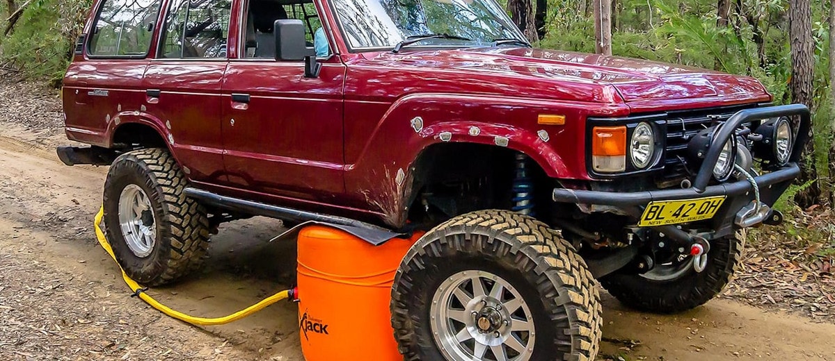 Big red truck with large wheels is parked on a dirt track. A bright orange exhaust jack under the front wheel with the hose leading to the tailpipe.