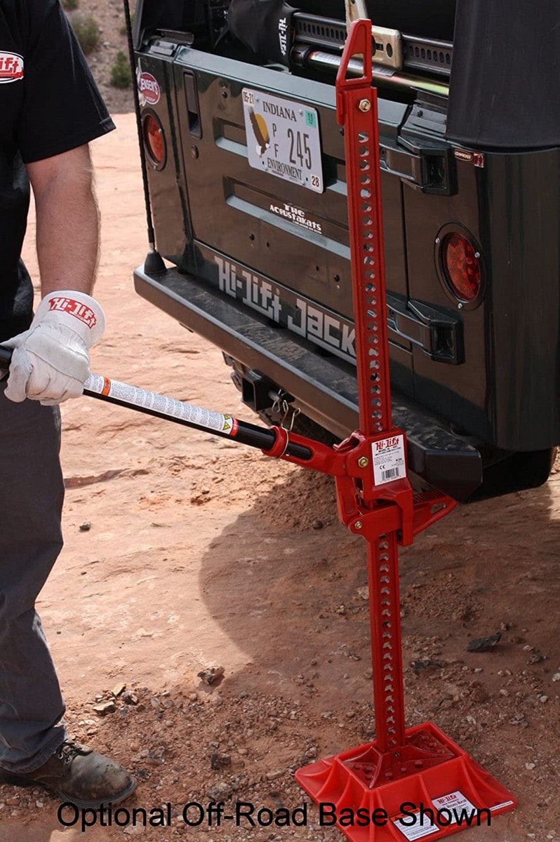 A red high lift jack is being used on the back of a black truck. A man with white gloves is pumping the lever.